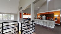 The River Oak Apartments clubhouse is a thoughtfully designed community space that impresses from the moment you enter. Soaring vaulted ceilings rise dramatically throughout, creating an expansive, airy atmosphere accentuated by an abundance of natural light pouring in through large multi-pane windows. Bold orange accent walls punctuate the neutral gray palette, lending the space a vibrant, modern energy.

At the heart of the clubhouse is a full kitchen and bar area outfitted with dark cabinetry, stainless steel appliances, and a sleek countertop bar with contemporary acrylic stools — ideal for entertaining and social gatherings. Adjacent to the kitchen, a comfortable lounge area features teal wingback chairs arranged around a large whiteboard wall, perfect for both relaxation and informal meetings.

The great room is anchored by a striking floor-to-ceiling fireplace flanked by generous windows that frame lush outdoor greenery, creating a warm and inviting focal point. A dedicated dining area with dark high-top tables and leather chairs provides an elegant spot for residents to gather, while vibrant abstract artwork adds a curated, gallery-like touch. The game room, complete with a ping pong table and casual seating, offers a fun and energetic retreat beneath its own vaulted ceiling. A glass-railed mezzanine overlooks the main level, tying the entire space together with architectural sophistication.