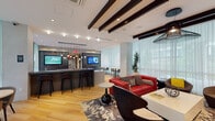 Located at 1100 Sixth St SW in Washington, DC, this contemporary amenity lounge is a well-appointed communal space designed for both relaxation and social engagement. The single-floor layout flows seamlessly across distinct zones, unified by warm hardwood flooring and a cohesive modern design aesthetic.

The main lounge area is anchored by bold red leather sofas accented with patterned throw pillows, arranged around a sleek white coffee table atop a graphic area rug. A wall-mounted flat-screen TV with a built-in fireplace surround serves as a focal point, while dramatic exposed dark wood ceiling beams add architectural character overhead. Striking pendant lighting, including sculptural wire and globe fixtures, provides ambient illumination throughout.

A bar counter with tall black stools offers a casual gathering spot, complemented by mounted digital displays and a textured decorative wall panel. Adjacent to this, a regulation billiards table sits beneath a bold cluster of industrial-style pendant lights, with large glass sliding doors opening directly to an outdoor patio area.

A dining nook features intimate round tables paired with curved upholstered chairs, bathed in soft natural light streaming through floor-to-ceiling sheer white curtains. These expansive windows provide views of the surrounding outdoor greenery, creating a bright and airy atmosphere throughout. Geometric three-dimensional wall tiling adds a refined design detail near the entryway, enhancing the overall contemporary character of the space.