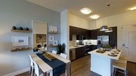 This single-floor residence showcases a thoughtfully designed open-concept layout that seamlessly connects the kitchen, dining, and living areas. The contemporary kitchen is a standout feature, outfitted with sleek dark cabinetry, stainless steel appliances including a refrigerator, range, microwave, and dishwasher, and a light stone backsplash that adds textural contrast. A kitchen island with a crisp white countertop provides additional prep space and serves as a natural dividing point between the kitchen and the rest of the living area. Warm hardwood-style flooring flows throughout, grounding the space with a sense of cohesion. The dining area comfortably accommodates a full-sized table with seating for multiple guests, accented by a large floor mirror and floating wood shelves that add depth and visual interest to the wall. Modern flush-mount ceiling lights and a stylish pendant fixture over the kitchen provide warm, even illumination throughout. A dedicated in-unit laundry area is conveniently tucked nearby, offering practical functionality without compromising the overall aesthetic. The neutral color palette of soft grays and warm whites creates a calm, inviting ambiance suited for both everyday living and entertaining.