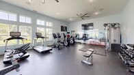 Located at 55 Dorset Crossing Drive in Simsbury, Connecticut, this fitness center offers a thoughtfully designed exercise space spread across a single floor. The room is characterized by impressively high ceilings that lend an open, airy feel, complemented by large windows along multiple walls that flood the interior with natural light and provide views of the surrounding greenery outdoors.

The light blue walls and durable rubber flooring create a clean, motivating atmosphere throughout. Cardio equipment is well-represented, with multiple treadmills, elliptical machines, and stationary and cycling bikes positioned to take advantage of the natural light. A dedicated free weights area features a multi-tiered dumbbell rack alongside a weight bench, while a cable/strength machine rounds out the resistance training options. Interlocking foam mat flooring defines the strength training zone, providing a safe surface for floor exercises.

A large wall-mounted flat-screen TV and a ceiling fan add comfort and convenience. Full-length mirrors along one wall help maximize the sense of space while supporting proper form during workouts. The fitness center is accessible via a set of glass-paneled double doors, and additional wall-mounted accessories such as resistance bands and foam rollers are also available. The overall design balances functionality with a bright, welcoming ambiance suitable for a wide range of fitness routines.