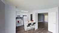 This well-designed two-floor residence offers a thoughtfully arranged layout that balances functionality and modern aesthetics. The main level showcases an open-concept floor plan where the kitchen flows naturally into the living area, creating a connected and airy atmosphere.

The kitchen is outfitted with crisp white cabinetry, white appliances including a refrigerator, electric range with overhead microwave, and ample counter space. A white breakfast bar extends from the kitchen and comfortably seats two, making it an ideal spot for casual dining or entertaining. The contrasting dark grey accent wall adds depth and visual interest to the space.

Durable wood-look flooring runs throughout the main living area, providing a warm and easy-to-maintain surface underfoot. The living room, visible from the kitchen, features neutral grey walls and light-filtering window treatments that allow natural light to brighten the space.

The clean white walls and ceilings throughout contribute to a bright, open feel, while the consistent flooring across rooms creates a cohesive visual flow. The two-floor layout offers additional living or sleeping space above, rounding out a practical and comfortable residence with a contemporary, move-in-ready appeal.