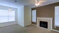 This two-story space offers a well-designed layout with comfortable and functional living areas. The main living room serves as a central gathering space, finished with wall-to-wall carpet that adds warmth underfoot. A fireplace with a clean white mantle anchors one wall, framed by a rich brown accent wall that creates a bold focal point against the otherwise neutral, light-toned walls. Crown molding runs along the ceiling perimeter, adding a refined architectural detail throughout the room. A ceiling fan with an integrated light fixture is centrally mounted, providing both air circulation and ambient lighting. Multiple windows fitted with horizontal blinds allow natural light to filter into the space, brightening the interior while maintaining privacy. The combination of neutral tones, classic molding details, and the warmth of the fireplace creates an inviting and comfortable ambiance. Spanning two floors, the property provides ample living space suited for a variety of lifestyle needs, with the overall design balancing practical functionality with approachable, traditional styling.