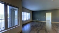 This single-floor space presents a thoughtfully designed interior that balances modern aesthetics with functional simplicity. The main living area immediately draws attention with its rich, dark hardwood flooring that runs throughout, adding warmth and a sense of depth to the space. Large windows fitted with horizontal blinds line one wall, flooding the interior with generous natural light and offering views of the surrounding outdoor scenery. The clean, smooth ceilings complement the open feel of the room, while neutral-toned walls provide a versatile backdrop for a variety of interior styles. A bold, deep teal-blue accent wall serves as a standout architectural feature, adding a layer of visual interest and character to the space. Baseboards in a warm wood tone frame the perimeter of the room, tying together the flooring and wall elements cohesively. Multiple electrical outlets are conveniently positioned throughout, reflecting practical, move-in-ready functionality. The overall ambiance is modern, airy, and understated, making it well-suited for a range of uses. The open layout encourages flexible furniture arrangements, while the interplay of natural light and rich finishes creates an inviting and polished environment throughout.