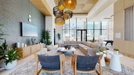 Located at 802 N 1st Ave in the heart of Phoenix, Arizona, this expansive single-floor amenity lounge offers a thoughtfully designed communal space spanning nearly 2,000 square feet. The interior is defined by soaring ceilings clad in warm wood paneling, large-format porcelain tile flooring, and dramatic floor-to-ceiling windows that flood the space with natural light and frame striking views of the Phoenix skyline.

The main lounge area anchors the space with a generous sectional sofa set on a raised wood platform, complemented by a textured accent wall and a wall-mounted display. An eclectic cluster of woven pendant lights adds warmth and visual interest overhead. Adjacent seating areas feature rope-woven accent chairs and intimate conversation groupings, ideal for socializing or relaxing.

A dedicated recreation zone includes a billiards table beneath modern pendant lighting, a wall-mounted TV, and built-in cue storage, providing a lively entertainment hub. A quieter alcove furnished with built-in shelving, a slatted wood partition, and a round coffee table offers a more private retreat for focused work or casual gatherings.

Natural greenery is woven throughout, enhancing the fresh, resort-inspired ambiance. Sheer floor-to-ceiling drapery softens the transition to outdoor views, while curated artwork and mixed-material wall treatments—including stone, tile mosaic, and wood—add layers of texture and sophistication throughout.