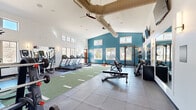 This dedicated fitness studio offers a thoughtfully organized workout space spread across a single open floor. The room is defined by a striking teal accent wall that contrasts with the surrounding white walls, creating an energetic and motivating atmosphere. Vaulted ceilings with exposed ductwork and a ceiling fan add a sense of vertical openness, while wall-mounted sconces provide warm, ambient lighting throughout.

The light wood-look flooring runs throughout the space, offering a clean and durable surface suited for a variety of exercise activities. Cardio equipment includes multiple stationary cycling bikes with integrated display screens and a rowing machine, all positioned to allow comfortable use and spacing. A multi-functional training rack with integrated weight storage, pull-up bars, and a heavy bag attachment anchors one side of the room for strength and functional training. A dumbbell rack stocked with a full range of weights provides additional resistance training options.

A wall-mounted flat-screen television serves as a focal point on the white end wall, ideal for guided workouts or entertainment. A dedicated yoga storage room is accessible via a separate door, keeping the main floor clear and organized. The space is efficiently ventilated with visible HVAC vents and benefits from natural light through a window near the exit door, completing a clean, functional, and well-appointed fitness environment.