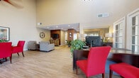This single-floor community or leasing office space makes a strong first impression with its dramatic vaulted ceilings and airy, open layout. Wood-look tile flooring flows seamlessly throughout, lending a clean and contemporary feel to every area of the space.

The reception area is anchored by a dark wood desk paired with bold red accent chairs, creating a professional yet inviting focal point. A wood-paneled media wall with a mounted television and cabinetry defines a comfortable lounge area furnished with patterned armchairs, offering a relaxed setting for visitors.

A dedicated seating area features two upholstered club chairs with vibrant yellow accent pillows, arranged alongside a round glass-top dining table surrounded by vivid crimson chairs — a colorful and energetic combination punctuated by framed abstract artwork on the warm beige walls. A large decorative clock adds a stylish finishing touch.

Multiple sets of French doors line the exterior walls, flooding the interior with abundant natural light and providing easy access to the surrounding outdoor areas. The high ceilings, enhanced by a ceiling fan and recessed lighting, contribute to the spacious and welcoming atmosphere throughout. Lush indoor plants are strategically placed to add warmth and a natural element to the overall design, completing the polished and functional character of this well-appointed space.