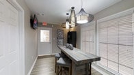 This single-floor space showcases a thoughtfully curated rustic-modern aesthetic that blends warmth and contemporary style seamlessly. The dining area serves as a focal point, centered around a reclaimed wood high-top table paired with upholstered bar-height chairs featuring nailhead trim detailing — a refined touch that elevates the overall design. Above, a striking trio of wire-frame pendant lights in varying sizes draws the eye upward, adding dramatic visual interest and serving as a true design statement against the textured ceiling. The walls are finished in a soft, warm gray tone that complements the light wood-look flooring, creating a cohesive and inviting atmosphere throughout. Natural light filters in generously through windows dressed with classic white plantation shutters, offering both brightness and privacy control. A wall-mounted flat-screen television provides a functional media element, while framed artwork adorns the walls, adding a curated, gallery-like quality to the space. A tall dark storage cabinet anchors one corner, offering practical storage without disrupting the visual flow. The space is well-lit, welcoming, and designed with both comfort and style in mind, making it well-suited for gathering and everyday use alike.