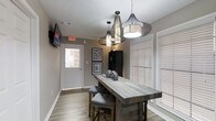 This single-floor space showcases a thoughtfully curated rustic-modern aesthetic that blends warmth and contemporary style seamlessly. The dining area serves as a focal point, centered around a reclaimed wood high-top table paired with upholstered bar-height chairs featuring nailhead trim detailing — a refined touch that elevates the overall design. Above, a striking trio of wire-frame pendant lights in varying sizes draws the eye upward, adding dramatic visual interest and serving as a true design statement against the textured ceiling. The walls are finished in a soft, warm gray tone that complements the light wood-look flooring, creating a cohesive and inviting atmosphere throughout. Natural light filters in generously through windows dressed with classic white plantation shutters, offering both brightness and privacy control. A wall-mounted flat-screen television provides a functional media element, while framed artwork adorns the walls, adding a curated, gallery-like quality to the space. A tall dark storage cabinet anchors one corner, offering practical storage without disrupting the visual flow. The space is well-lit, welcoming, and designed with both comfort and style in mind, making it well-suited for gathering and everyday use alike.
