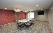 This thoughtfully designed conference space makes a strong impression through a compelling blend of rustic elegance and contemporary style. The room's most striking feature is a vivid, textured accent wall in warm shades of red, orange, and gold, which adds dramatic visual depth and energy to the space. Complementing this bold design choice are two statement antler chandeliers suspended from the ceiling, lending a distinctive, nature-inspired character to the overall aesthetic.

The main conference area centers around a large, sleek white table surrounded by colorful, multicolored patchwork upholstered rolling chairs, offering both comfort and a playful pop of color. A wall-mounted display screen provides essential presentation capabilities, while a window along one wall invites natural light into the space.

A separate, more intimate seating arrangement with a round glass table and matching chairs provides a flexible alternative for smaller discussions or informal meetings. The space is partially enclosed by floor-to-ceiling glass partition walls with black framing, offering visual connectivity while maintaining a sense of privacy.

Light gray wood-look flooring runs throughout, tying the various elements together with a clean, modern foundation. Recessed lighting and exposed concrete structural columns add further architectural interest, making this a highly functional yet visually engaging professional environment.