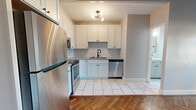 This thoughtfully designed single-floor unit makes excellent use of its compact footprint, offering a living room, kitchen, and bathroom with a cohesive, modern aesthetic. The living area is defined by rich, herringbone-patterned hardwood floors that lend a timeless, elegant character to the space. A ceiling fan and a window allowing natural light complete the inviting atmosphere, while neutral gray walls create a clean, versatile backdrop.

The kitchen is a standout feature, showcasing a thorough renovation with crisp white shaker cabinetry, a classic subway tile backsplash, and marble-look porcelain tile flooring. A full suite of stainless steel appliances — including a refrigerator, range, microwave, and dishwasher — is complemented by a matte black faucet and a modern globe chandelier, blending contemporary and traditional design elements seamlessly.

The bathroom continues the refined aesthetic with floor-to-ceiling white subway tile lining a glass-enclosed walk-in shower, accented by bold matte black hardware and fixtures. A window within the shower brings in natural light, adding an airy quality to the space. Every room reflects careful attention to detail, resulting in a polished, move-in-ready unit that balances classic charm with modern upgrades.