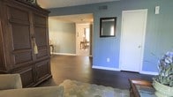 This single-story home presents a well-connected layout with a comfortable, cohesive aesthetic throughout. The living area showcases rich, dark hardwood flooring that extends seamlessly into adjacent rooms, creating a unified and grounded feel underfoot. Walls are painted in a soft blue-gray tone, lending the interior a calm, consistent atmosphere from room to room.

The main living space is anchored by a substantial dark wood armoire, adding a traditional character to the room. A decorative mirror in a dark frame is mounted on the accent wall, complementing the warm wood tones present in the furnishings. A patterned area rug in muted greens introduces a subtle contrast against the dark flooring.

An open passageway connects the living area to a secondary room, which transitions into what appears to be a dining area. The dining space, visible through the passage, features lighter flooring and traditional wooden furniture, including a classic table and chairs, with natural light filtering in through a window. Ceiling-mounted HVAC vents indicate central climate control throughout. The overall ambiance is traditional and lived-in, with a warm, approachable character.