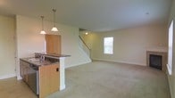 This two-floor residence offers a well-designed layout that balances functionality and comfort. The main level showcases an open-concept floor plan, seamlessly connecting the kitchen and living area into one cohesive space. The kitchen features warm wood cabinetry, a dark granite-style countertop with a built-in sink, and a peninsula that serves as both a prep area and a casual dining spot. Pendant lighting with classic globe fixtures hangs above the peninsula, providing focused illumination and a clean, understated aesthetic. The adjacent living area is carpeted in neutral tones, creating a soft and inviting atmosphere. A window on the main level allows natural light to filter in, brightening the open space. A traditional staircase with white-painted balusters and a wood handrail anchors the far end of the main floor, leading to the second level. Smooth, light-colored walls and white baseboards run throughout, contributing to a crisp and clean interior. The overall ambiance is straightforward and move-in ready, with a practical layout that maximizes usable space across both floors.