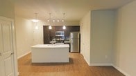 This single-floor space is anchored by a well-appointed open-concept kitchen that seamlessly connects to the adjacent living area, creating a cohesive and functional layout. The kitchen features dark espresso cabinetry contrasted by a sleek white kitchen island with an integrated sink, offering both prep space and a casual gathering spot. Stainless steel appliances, including a refrigerator, gas range, and built-in microwave, provide a modern, polished aesthetic. A tile backsplash in cool blue tones adds a subtle decorative accent behind the range. Pendant lighting hangs above the island, while a track lighting system illuminates the main kitchen workspace, ensuring the area is well-lit. Warm hardwood-style flooring extends throughout the open space, adding continuity and a welcoming feel. The open layout flows into a spacious living area flanked by neutral-toned walls, offering versatility for various furniture arrangements. The overall ambiance is contemporary and clean, with a balance of dark and light finishes that create a modern, inviting atmosphere well-suited for everyday living and entertaining.