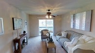 This single-floor residential space offers a warm and inviting atmosphere, thoughtfully designed for comfortable everyday living. The living room serves as the centerpiece of the home, featuring soft wall-to-wall carpeting that adds a cozy underfoot feel throughout. A ceiling fan with an integrated light fixture hangs prominently overhead, providing both air circulation and ambient lighting that casts a pleasant glow across the room. Natural light floods the space through a large sliding glass door, which opens onto a private balcony offering views of the surrounding exterior — a standout feature that enhances the overall sense of openness. The neutral, warm-toned walls create a versatile backdrop that complements a variety of décor styles. The living area is well-proportioned, comfortably accommodating a full sofa, accent chair, and coffee table arrangement, making it ideal for both relaxing and entertaining. A console table along one wall provides additional display and storage space. The sliding glass door not only brings in an abundance of natural light but also creates a seamless visual connection between the interior and the outdoor balcony, contributing to the bright and airy character of this well-appointed single-floor home.