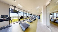 This contemporary fitness center offers a thoughtfully designed, single-level workout space that provides a comprehensive exercise environment. The facility is defined by an expansive wall of floor-to-ceiling windows running the length of the room, flooding the interior with natural light and offering sweeping panoramic views of the surrounding landscape — an inspiring backdrop for any workout.

The cardio zone is prominently featured along the window wall and includes a row of treadmills, elliptical trainers, and stationary bikes, each positioned to take full advantage of the outdoor views. Multiple wall-mounted flat-screen televisions are strategically placed throughout for entertainment during workouts.

A dedicated strength and functional training area occupies the opposite end of the space, equipped with a multi-station cable machine, a medicine ball rack stocked with various weighted balls, and kettlebells. A full-length mirror panel along one wall enhances the sense of space and supports proper form during exercise.

The interior design is vibrant and energizing, highlighted by a striking large-scale botanical wall mural rendered in gold, grey, and white tones. Accent walls in a bright yellow-green complement the light-toned flooring, creating a lively and motivating atmosphere. A glass-enclosed entry and recessed ceiling lighting complete the polished, modern aesthetic of this well-appointed fitness facility.