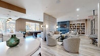 Located at 15 Washington Ave in Suffern, NY, this sophisticated single-floor lounge space offers a thoughtfully curated amenity experience with a contemporary design aesthetic. Light-toned hardwood flooring runs throughout, complementing a neutral palette of warm grays, taupes, and deep navy accents.

The open-concept layout is artfully divided by a striking floor-to-ceiling slatted wood screen partition, creating distinct yet visually connected zones. A central lounge area features plush swivel armchairs and a sectional sofa arranged around a glass coffee table, anchored by a bold navy area rug with gold accents. Opposite, a dramatic blue tile-clad fireplace with a wall-mounted display above serves as a focal point, flanked by built-in open shelving with curated décor.

A game area houses a billiards table set beneath globe pendant lights, bathed in natural light from oversized windows overlooking the outdoors. A casual dining nook with high-top seating and bistro tables adds a social gathering space. Textured wallcovering with a subtle grid pattern adds visual depth to key walls, while recessed lighting throughout ensures a warm, inviting ambiance. The space also features a wet bar area with sleek cabinetry, completing this well-appointed communal retreat.
