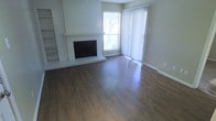 This single-story space centers around a spacious living area finished with warm, rich hardwood flooring that extends throughout the room. A prominent white brick fireplace serves as the focal point of the space, flanked by built-in shelving that offers both display and storage opportunities. The fireplace surround is finished in white-painted brick, lending a clean, classic aesthetic to the room.

Natural light fills the space generously through a large window and an adjacent floor-to-ceiling sliding glass door dressed with vertical blinds, which together create a bright and airy atmosphere. The sliding door provides access to an outdoor area and frames a view of mature trees beyond. Neutral-toned walls complement the flooring and fireplace, creating a cohesive, understated palette throughout.

The open layout of the living area offers a versatile arrangement, accommodating a variety of furniture configurations. The combination of the hardwood floors, white brick detailing, and abundant natural light gives the space a welcoming and functional character suitable for everyday living.