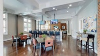 This single-floor community amenity space is thoughtfully designed to accommodate a variety of social and functional activities. Rich hardwood flooring flows throughout, anchoring a cohesive and inviting atmosphere across every zone.

The main gathering area combines casual dining and lounge seating in an open-plan layout. Mixed seating arrangements — including bar-height tables with upholstered chairs and comfortable sofas with accent armchairs — cater to both lively gatherings and relaxed conversation. Multiple wall-mounted flat-screen TVs are suspended from a coffered ceiling with recessed lighting, keeping residents connected and entertained. Large windows along the perimeter flood the space with abundant natural light and offer views of the outdoors.

Exposed brick accent walls add warmth and industrial character throughout, complementing the contemporary furnishings. A dedicated billiards room features a full-size pool table set against a striking brick wall with its own mounted TV and additional seating.

The fully equipped kitchen is a standout feature, boasting granite countertops, dark cabinetry, stainless steel appliances, and ample prep space — ideal for hosting events. A glass-enclosed private meeting or lounge room provides a quieter retreat within the larger space. High-mounted pendant lighting and curated artwork complete the polished, community-focused aesthetic.