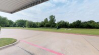 Located in Frisco, this single-level outdoor space offers a well-maintained environment characterized by generous open areas and natural surroundings. The property features a expansive fenced yard enclosed by sturdy black metal railings, providing a secure and clearly defined boundary. The lush green lawn stretches broadly, framed by a dense backdrop of mature deciduous trees that offer shade and a sense of privacy from the surrounding landscape.

A covered paved area is situated adjacent to the yard, marked with painted lines and sheltered by an overhead canopy structure — suitable for vehicle parking or covered outdoor activities. The transition between the hardscaped and landscaped areas is clean and well-defined, with a raised curb separating the concrete surface from the grassy grounds.

Decorative natural boulders are placed within the yard, adding subtle landscaping detail to the otherwise open green space. The black iron perimeter fencing is consistent throughout, maintaining a cohesive and secure aesthetic. The overall ambiance is open, green, and functional, with a park-like quality enhanced by the tree line and well-kept turf — creating a versatile outdoor space suited for a variety of uses.