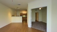This single-story space offers an open-concept layout that connects the main living area to the kitchen and additional rooms. The living area features warm hardwood-style flooring, white walls with crown molding, and a track lighting fixture that provides ample illumination. A raised breakfast bar separates the living space from the kitchen, which is equipped with warm-toned wood cabinetry, dark countertops, and under-cabinet lighting that adds a functional ambiance. The kitchen benefits from recessed lighting, enhancing the workspace visibility. An adjoining room, accessible through a wide doorway, transitions to plush carpeted flooring and is fitted with a ceiling fan with a light fixture, offering both comfort and airflow. A hallway visible from this secondary room leads further into the unit, suggesting additional private spaces beyond. HVAC vents are positioned throughout the ceiling, ensuring consistent climate control across the layout. The overall design balances practicality and comfort, with a neutral color palette and warm finishes creating a cohesive and welcoming atmosphere throughout the space.