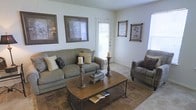 This single-floor space presents a traditionally styled living room that balances comfort and classic design. The living room is anchored by a generously sized upholstered sofa in muted earth tones, complemented by a matching armchair, both adorned with decorative throw pillows in coordinating patterns. A rectangular wooden coffee table with a metal frame serves as the centerpiece, styled with tall candlestick holders and a decorative keepsake box, adding a curated, homey touch. The flooring features neutral carpeting partially covered by a patterned area rug that adds warmth and visual interest to the space. Natural light enters through multiple windows fitted with white blinds, as well as through a glass-paneled door that provides an additional source of brightness and contributes to an open, airy feel. The walls are painted in a soft, light neutral tone and are decorated with several framed artwork pieces in warm, coordinating tones. The textured ceiling adds subtle architectural character. Overall, the living area conveys a welcoming and well-appointed atmosphere suited to relaxed, everyday living.