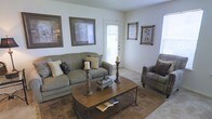 This single-floor space presents a traditionally styled living room that balances comfort and classic design. The living room is anchored by a generously sized upholstered sofa in muted earth tones, complemented by a matching armchair, both adorned with decorative throw pillows in coordinating patterns. A rectangular wooden coffee table with a metal frame serves as the centerpiece, styled with tall candlestick holders and a decorative keepsake box, adding a curated, homey touch. The flooring features neutral carpeting partially covered by a patterned area rug that adds warmth and visual interest to the space. Natural light enters through multiple windows fitted with white blinds, as well as through a glass-paneled door that provides an additional source of brightness and contributes to an open, airy feel. The walls are painted in a soft, light neutral tone and are decorated with several framed artwork pieces in warm, coordinating tones. The textured ceiling adds subtle architectural character. Overall, the living area conveys a welcoming and well-appointed atmosphere suited to relaxed, everyday living.