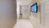 This mailroom is designed with both functionality and modern aesthetics in mind. The space is laid out as a corridor-style room, providing ample wall space to accommodate multiple banks of individual metal mailboxes lining the walls. The mailboxes are arranged in a grid format across several panels, offering organized mail access for a large number of residents or occupants.

The flooring throughout is polished white ceramic tile, lending the space a clean, bright appearance. One of the most distinctive design elements is the textured stone-finish accent wall in a warm taupe-gray tone, which runs the length of one side of the room, adding visual depth and contrast against the crisp white walls.

Recessed lighting in the white ceiling provides even, well-distributed illumination throughout the corridor. A wall-mounted flat-screen display is positioned prominently, offering informational or community content. A stainless steel recycling bin is conveniently located near the mailboxes, encouraging responsible waste disposal. The overall ambiance is clean, contemporary, and efficiently organized, reflecting a well-maintained communal service area.