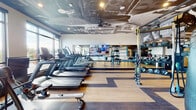 This contemporary fitness center is designed to deliver a premium workout experience, combining thoughtful architecture with a comprehensive range of equipment. The space is unified by striking, artistic ceiling murals — an abstract, large-scale graphic design that adds a bold visual identity and sets the gym apart from conventional fitness facilities. The flooring throughout features a warm wood-tone finish with contrasting dark stripe inlays, lending a polished, dynamic aesthetic to the open layout.

The cardio zone is well-appointed with a row of treadmills and rowing machines, positioned alongside floor-to-ceiling windows that flood the area with natural light and offer open outdoor views. A large flat-screen television is mounted centrally on a mirrored wall, enhancing the workout atmosphere.

The strength and functional training area includes a multi-functional cable and resistance station, a punching bag for boxing training, elliptical machines, and an organized rack of kettlebells and dumbbells. Full-length mirrors along multiple walls amplify the sense of space while providing visual feedback during training.

A dedicated accessory wall features neatly organized storage racks stocked with medicine balls, foam rollers, resistance bands, balance discs, and other functional training tools. Ceiling fans and recessed lighting ensure comfort and visibility throughout. The overall ambiance is energetic yet refined, making this fitness center both functional and visually impressive.