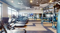 This contemporary fitness center is designed to deliver a premium workout experience, combining thoughtful architecture with a comprehensive range of equipment. The space is unified by striking, artistic ceiling murals — an abstract, large-scale graphic design that adds a bold visual identity and sets the gym apart from conventional fitness facilities. The flooring throughout features a warm wood-tone finish with contrasting dark stripe inlays, lending a polished, dynamic aesthetic to the open layout.

The cardio zone is well-appointed with a row of treadmills and rowing machines, positioned alongside floor-to-ceiling windows that flood the area with natural light and offer open outdoor views. A large flat-screen television is mounted centrally on a mirrored wall, enhancing the workout atmosphere.

The strength and functional training area includes a multi-functional cable and resistance station, a punching bag for boxing training, elliptical machines, and an organized rack of kettlebells and dumbbells. Full-length mirrors along multiple walls amplify the sense of space while providing visual feedback during training.

A dedicated accessory wall features neatly organized storage racks stocked with medicine balls, foam rollers, resistance bands, balance discs, and other functional training tools. Ceiling fans and recessed lighting ensure comfort and visibility throughout. The overall ambiance is energetic yet refined, making this fitness center both functional and visually impressive.