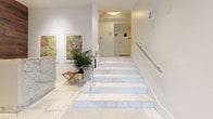 This building lobby, identified as "The Bond," presents a carefully designed entry experience that balances functionality with refined aesthetics. The space is anchored by striking white marble flooring with subtle grey veining, which flows continuously throughout the main corridor, staircase, and entry vestibule, establishing a cohesive and upscale visual identity.

Upon entry through the solid wood-framed glass doors, visitors are greeted by a sleek marble concierge desk set against a feature wall clad in rich, dark wood planking. This warm tonal contrast between the white marble and deep wood tones defines the lobby's design language. Recessed ceiling lighting, combined with flush-mount fixtures, ensures the space is evenly and cleanly illuminated.

The lobby extends into a well-organized mail corridor lined with individual resident mailboxes, accompanied by a decorative round mirror and wall sconce. A set of marble stairs with polished chrome railings leads to the upper level, flanked by framed artwork and a potted plant that introduce organic warmth. The overall ambiance is clean, contemporary, and professionally appointed, reflecting a high standard of building design.