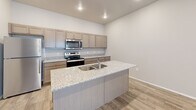 This single-floor unit offers a well-organized layout comprising a combined kitchen and living area, one bedroom, and a dedicated mudroom entry. The kitchen serves as a central feature, equipped with stainless steel appliances including a refrigerator, electric range, and over-the-range microwave. Taupe shaker-style cabinetry pairs with granite-patterned countertops, and a large center island with a double basin sink provides additional workspace and a natural gathering point. Light wood-look flooring runs throughout the kitchen and living area, contributing to a clean, contemporary aesthetic. The living room is bright and open, featuring a ceiling fan with integrated lighting, exposed cylindrical ductwork that adds an industrial touch, and a full sliding glass door dressed with vertical blinds to allow natural light. The bedroom offers carpeted flooring, a ceiling fan, a window for natural ventilation, and a wood-toned door consistent with the unit's design language. A dedicated mudroom near the entry provides a built-in bench with storage cubbies and a row of wall hooks, offering practical organization space. Recessed lighting and a neutral gray-white color palette unify the space throughout, creating a modern and functional environment.