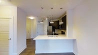 This 1-bedroom, 1-bathroom unit (A2 floor plan) is thoughtfully designed with an open-concept layout that connects the kitchen and living areas seamlessly. The kitchen serves as the focal point, featuring a large peninsula with a crisp white countertop and an integrated sink, ideal for both meal preparation and casual dining. Three brushed nickel pendant lights hang above the peninsula, providing targeted task lighting while adding a contemporary aesthetic touch.

Dark espresso cabinetry lines the kitchen walls, offering generous storage and creating a striking contrast against the light countertops and neutral walls. A full suite of stainless steel appliances, including a French door refrigerator with ice and water dispenser, microwave, and range, equips the kitchen for everyday use. A neutral tile backsplash adds texture and visual interest behind the appliances.

The flooring throughout is rich, dark hardwood-style planking, lending warmth and continuity across the open living space. Additional track lighting in the main area ensures the space is well-lit. Multiple interior doors provide access to the bedroom, bathroom, and entryway, completing this efficiently designed, move-in-ready unit.