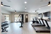 Located at 4781 SW 36 Rd in Gainesville, Florida, this well-appointed fitness center offers a thoughtfully designed single-floor layout with a comfortable, open workout environment. The space features light wood-style flooring, crisp white walls, and recessed lighting complemented by multiple ceiling fans that promote airflow throughout the facility.

The cardio zone is equipped with a lineup of True-brand treadmills, an elliptical trainer, and a stationary bike, each positioned in front of wall-mounted flat-screen televisions for an engaging workout experience. A dedicated strength training area includes a multi-station cable machine and a full dumbbell rack with mirrored walls, providing a comprehensive free-weight training setup.

Natural light fills the space through several windows with roller shades, offering views of what appears to be an outdoor pool and patio area. Additional wall art and framed prints add a polished, welcoming aesthetic to the interior. The facility also includes supplementary rooms, likely serving as storage or utility spaces. Overall, the fitness center presents a clean, modern atmosphere well-suited for a full range of cardio and strength training activities.