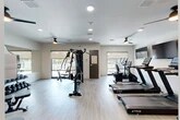Located at 4781 SW 36 Rd in Gainesville, Florida, this well-appointed fitness center offers a thoughtfully designed single-floor layout with a comfortable, open workout environment. The space features light wood-style flooring, crisp white walls, and recessed lighting complemented by multiple ceiling fans that promote airflow throughout the facility.

The cardio zone is equipped with a lineup of True-brand treadmills, an elliptical trainer, and a stationary bike, each positioned in front of wall-mounted flat-screen televisions for an engaging workout experience. A dedicated strength training area includes a multi-station cable machine and a full dumbbell rack with mirrored walls, providing a comprehensive free-weight training setup.

Natural light fills the space through several windows with roller shades, offering views of what appears to be an outdoor pool and patio area. Additional wall art and framed prints add a polished, welcoming aesthetic to the interior. The facility also includes supplementary rooms, likely serving as storage or utility spaces. Overall, the fitness center presents a clean, modern atmosphere well-suited for a full range of cardio and strength training activities.