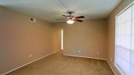 This single-story space presents a clean, functional layout centered around a spacious carpeted room. The room features warm beige walls that lend a neutral, welcoming atmosphere, complemented by light-toned carpet flooring that extends throughout. A five-blade ceiling fan with an integrated light fixture serves as the central overhead feature, providing both ventilation and ambient lighting. The white ceiling offers a crisp contrast to the warm wall tones, and a ceiling-mounted HVAC vent indicates a centralized climate control system. White baseboards frame the perimeter of the room, adding a subtle architectural detail. Electrical outlets are positioned along the walls at standard intervals, and a doorway on one wall leads to an adjacent room or hallway, suggesting a connected layout. The overall ambiance is straightforward and move-in ready, offering a blank canvas well-suited for a variety of furnishing arrangements. The simple, uncluttered design emphasizes practicality and ease of use throughout the space.