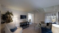 This single-floor residence offers a thoughtfully arranged living area characterized by its angled, beam-accented ceiling, which adds architectural interest and a sense of depth to the space. The living room is anchored by a wall-mounted flat-screen television above a sleek black console table, which provides both display and storage space. Neutral-toned carpeting runs throughout, complementing the light gray walls and contributing to a calm, cohesive aesthetic.

Seating is arranged around a glass-top coffee table, creating a comfortable and functional gathering area. A floor lamp provides ambient lighting, while a window dressed with vertical blinds allows natural light to filter in. An in-wall air conditioning unit is also present, offering climate control convenience.

A defined entryway with a paneled white door connects to an adjoining dining area, visible through an open passage. The dining space features a chair and table arrangement beneath a ceiling fan, and a large window with sheer coverings enhances brightness. The open flow between the living and dining areas reinforces a spacious, connected feel throughout the home.
