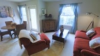 This single-floor living space centers around a comfortable living room designed with a warm, casual aesthetic. The room is furnished with plush red upholstered seating, including a generously sized armchair adorned with a soft throw blanket and decorative pillows, complementing a matching sofa. A dark wood coffee table with a metal frame serves as the room's focal point, topped with a decorative zebra-print vase that adds a subtle stylistic accent. Natural light fills the space through two large windows dressed in blue patterned curtains, offering views of surrounding greenery outside. A glass-paneled door on the adjacent wall provides additional light and likely leads to an outdoor area, further enhancing the bright, airy feel of the room. Neutral carpeting throughout grounds the space with a soft, understated base. A rustic wooden cabinet positioned in the corner adds storage while contributing to the room's warm, traditional character, displaying decorative figurines on top. Overall, the living area presents a relaxed and inviting atmosphere, thoughtfully arranged for both comfort and everyday functionality.