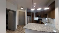 This single-floor space showcases a thoughtfully designed open-concept layout that seamlessly connects the kitchen and living areas. The kitchen is a standout feature, highlighted by sleek granite countertops with a classic speckled finish and rich dark wood cabinetry that extends to the upper walls, providing generous storage. Stainless steel appliances, including a refrigerator, microwave, and range, lend a contemporary, polished look to the space. A kitchen island with pendant lighting above serves as both a functional workspace and an inviting gathering spot. Track lighting complements the pendant fixtures, ensuring the kitchen is well-lit throughout. The flooring throughout the unit features warm-toned wood-look planks that add a sense of continuity and warmth to the overall aesthetic. A white subway tile backsplash adds a clean, timeless contrast to the darker cabinetry. The entryway is defined by a sturdy, modern door with clean grey trim, contributing to the unit's contemporary character. The white walls and ceilings keep the interior feeling bright and open, while the efficient use of space throughout speaks to a well-considered, functional floor plan ideal for everyday living.