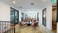 Located at 1464 Laurel Sound Ln in Sanford, Florida, this single-floor commercial space offers a thoughtfully designed layout that balances open collaboration with private functionality. The interior is defined by a clean, contemporary aesthetic, featuring crisp white walls, recessed lighting, and lush green carpet flooring that flows seamlessly throughout. Large black-framed windows invite generous amounts of natural light, offering views of the surrounding Florida landscape including palm trees.

A welcoming central lounge area serves as the heart of the space, furnished with rust-toned armchairs and a round coffee table — ideal for informal gatherings or casual meetings. Multiple private offices or meeting rooms are accessible via striking dark-framed glass doors, providing visual transparency while maintaining acoustic separation. One room features a sleek conference table with contemporary white chairs, suited for professional meetings.

A dedicated workstation area offers a built-in countertop desk with bar-height seating, a printer, and direct window views — a practical space for day-to-day office tasks. Additional lounge seating nooks are distributed throughout, each accented with decorative greenery and tasteful artwork. A staircase with a black metal and wood railing connects the floor to other levels of the building. The overall ambiance is modern, professional, and inviting.