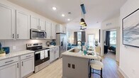 Located in Pensacola, FL, this well-appointed one-bedroom, one-bathroom residence offers an efficient and modern single-floor layout designed to maximize comfort and functionality. The open-concept kitchen flows seamlessly into the living area, creating a connected and airy atmosphere. The kitchen is outfitted with gray shaker-style cabinetry, granite countertops, a full suite of stainless steel appliances including a refrigerator with ice maker, electric range, and microwave, and a central island with a built-in sink and bar seating beneath sleek pendant lighting. Light wood-style flooring extends through the kitchen and living areas, enhancing the sense of openness. The living room features natural light from multiple windows with drape treatments and a ceiling fan overhead. The bedroom offers plush carpeting, a ceiling fan, and a window with exterior views, connected to a walk-in closet with wire shelving systems providing ample storage. The full bathroom includes light wood-style flooring, a soaking tub with tile surround, a granite-topped vanity, and direct access to the bedroom. Recessed lighting and a clean, neutral palette tie the entire space together with a modern, cohesive aesthetic.