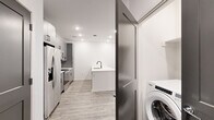 This thoughtfully designed one-bedroom, one-bathroom unit offers a modern aesthetic with clean lines and contemporary finishes throughout its single-floor layout. The open-concept kitchen and living area serves as the heart of the space, anchored by a striking white waterfall island with an undermount sink and matte black faucet. The kitchen features gray shaker-style cabinetry with black hardware, white quartz countertops, a geometric tile backsplash, and a full suite of stainless steel appliances. Light wood-look flooring runs throughout the main living area, complementing the bright white walls and recessed lighting. The living area benefits from natural light through large windows fitted with blinds, and a ceiling fan overhead. A dedicated in-unit washer is conveniently tucked behind a paneled door near the kitchen. The generously sized bedroom is finished with plush carpet, a ceiling fan, a window for natural light, and dark two-panel doors leading to a well-organized walk-in closet with built-in shelving. The bathroom continues the modern theme with a gray vanity, white quartz countertop, an illuminated LED mirror, subway-tiled tub surround, and matte black fixtures throughout.