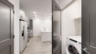 This thoughtfully designed one-bedroom, one-bathroom unit offers a modern aesthetic with clean lines and contemporary finishes throughout its single-floor layout. The open-concept kitchen and living area serves as the heart of the space, anchored by a striking white waterfall island with an undermount sink and matte black faucet. The kitchen features gray shaker-style cabinetry with black hardware, white quartz countertops, a geometric tile backsplash, and a full suite of stainless steel appliances. Light wood-look flooring runs throughout the main living area, complementing the bright white walls and recessed lighting. The living area benefits from natural light through large windows fitted with blinds, and a ceiling fan overhead. A dedicated in-unit washer is conveniently tucked behind a paneled door near the kitchen. The generously sized bedroom is finished with plush carpet, a ceiling fan, a window for natural light, and dark two-panel doors leading to a well-organized walk-in closet with built-in shelving. The bathroom continues the modern theme with a gray vanity, white quartz countertop, an illuminated LED mirror, subway-tiled tub surround, and matte black fixtures throughout.