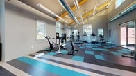 This fitness center is a thoughtfully designed, single-floor facility offering a comprehensive range of workout options. The space is defined by its striking architectural character, featuring soaring vaulted ceilings with exposed yellow structural beams and industrial-style ductwork that lend an open, modern aesthetic. Large windows throughout the facility flood the interior with natural light and provide views of the surrounding greenery and neighborhood.

The flooring consists of a bold checkerboard and striped pattern in teal, gray, and white rubber tiles, giving the space a vibrant and energetic feel. The main workout area is generously proportioned, accommodating a variety of equipment including spin bikes, elliptical trainers, treadmills, a weight bench, and a full dumbbell rack with a broad range of free weights. A functional cable training machine is also available, positioned on its own designated mat area.

A large wall-mounted display screen serves as a focal point in the main exercise zone, complemented by mirrored walls that enhance the sense of depth and space. Glass-paneled walls and doors reveal an adjacent racquetball court, adding a dynamic sporting dimension to the facility. A stationary bicycle is also visible near the entrance area. Overall, the fitness center delivers a bright, spacious, and well-appointed environment suited for a wide variety of training activities.