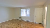 This single-floor space offers a straightforward and practical layout highlighted by warm hardwood flooring that runs throughout the main living area. The room is bathed in natural light streaming through a large, centrally positioned window that overlooks an exterior brick structure and railing, providing a view of the surrounding building environment. The white-painted walls and white ceiling contribute to a bright, open atmosphere, making the space feel airy despite its modest footprint. A wall-mounted air conditioning unit sits beneath the window, complemented by baseboard heating that runs along the perimeter of the room, ensuring year-round climate comfort. A white paneled door on the right side of the space provides entry or access to an adjacent area. The overall aesthetic is clean and understated, with neutral tones throughout that offer a versatile canvas for customization. The space presents a functional, no-frills environment well-suited for those seeking a straightforward living or working area with essential amenities already in place.