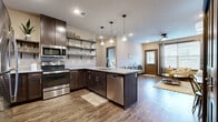 This single-floor residence offers a thoughtfully designed open-concept layout that seamlessly connects the kitchen, dining, and living areas. The kitchen is a standout feature, showcasing rich dark espresso cabinetry, granite countertops, stainless steel appliances including a refrigerator, electric range, microwave, and dishwasher, as well as open floating shelves that add a modern, airy touch. A peninsula with a sink provides additional workspace and casual seating. Pendant lighting illuminates the kitchen, while a ceiling fan keeps the living area comfortable. Warm-toned wood-look flooring runs throughout the main living spaces, complementing the neutral wall tones and creating a cohesive, inviting ambiance. The living room offers a relaxed, comfortable atmosphere with recessed lighting and access to a front entry door flanked by windows that bring in natural light. The bedroom is carpeted for warmth and comfort, offering direct sightlines to the kitchen and featuring a dedicated walk-in closet complete with wire shelving for ample storage. The bathroom is clean and functional, equipped with a granite vanity, dark wood cabinetry, a full tub-shower combination, and wood-look flooring. A laundry area with a stacked washer and dryer is also included within the unit for added convenience.