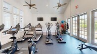 Located at 22052 Stratford Way in Athens, Alabama, this dedicated exercise room delivers a comprehensive fitness experience within a bright, thoughtfully designed space. The room's most striking architectural feature is its soaring vaulted ceiling, which creates an open, airy atmosphere and is complemented by two ceiling fans that ensure steady airflow during workouts. Recessed lighting and large French-style glass doors and windows flood the interior with natural light, enhancing the clean, white walls and contributing to an energizing environment. The flooring transitions between warm hardwood and durable dark rubber gym matting, providing both aesthetic appeal and functional support for exercise equipment. The room is outfitted with an impressive array of cardio machines, including multiple treadmills, ellipticals, and stationary bikes, as well as a cable strength-training machine. A well-organized rack along one wall holds a variety of exercise balls, resistance bands, and other accessories. Two wall-mounted flat-screen televisions provide entertainment during workouts. Additional doors lead to adjacent utility areas, adding convenience to the space. Overall, this fitness room offers a professional-grade workout environment with a welcoming, light-filled ambiance.