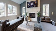 This single-floor clubhouse-style space is designed with a contemporary aesthetic, blending comfort and sophistication across several distinct yet open zones. The main gathering area showcases soaring ceilings and herringbone-patterned wood-look flooring, lending a refined, upscale character to the space. A full-sized pool table anchors the game area, positioned beneath a striking gallery wall featuring framed feather artwork and bold medallion-style wall pieces.

Adjacent lounge areas offer versatile seating arrangements, including plush sofas, accent chairs, and coffee tables styled with decorative accessories. One seating nook centers on a stacked-stone fireplace with a wall-mounted television above, flanked by tall windows that allow natural light to fill the room and provide exterior views. The cool-toned charcoal accent walls in this area contrast elegantly with the light stone surround.

A separate sitting area features a row of geometric diamond-framed mirrors reflecting additional natural light from large windows. A chandelier-lit anteroom is visible through an arched passage, adding architectural interest. Recessed lighting throughout ensures the space is well-illuminated, while curated greenery and tasteful décor contribute to a welcoming, polished ambiance.