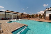 This amenity space is a well-appointed outdoor pool area designed to deliver a resort-like experience. At its center is a generously sized swimming pool with crystal-clear water, featuring defined entry steps and a shallow wading section alongside a deeper area, making it accessible for a wide range of users. The pool is surrounded by an expansive brick paver deck, lined with rows of modern lounge chairs and orange-accented umbrellas that provide ample space for sunbathing and relaxation under open skies.

Adjacent to the pool, a covered cabana structure serves as the architectural anchor of the space. Built in a clean, contemporary style with white board-and-batten siding and dark metal accents, it features an open-air lounge area outfitted with teak furniture, plush cushions, patterned ottomans, and blue area rugs. Ceiling fans keep the space comfortable, while privacy curtains can be drawn for a more intimate setting. The structure also features large, floor-to-ceiling sliding glass doors that connect the interior to the pool deck seamlessly.

Lush mature trees frame the perimeter of the space, providing a natural backdrop and a sense of privacy. The overall ambiance is bright, airy, and inviting, reflecting a thoughtfully designed outdoor retreat suited for both leisure and social gatherings.