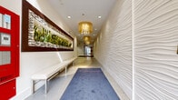 The Avanath+ lobby is a thoughtfully designed single-floor entrance space that combines contemporary aesthetics with functional elements to create a polished and welcoming environment. Upon entry, visitors are greeted by glass-paneled doors that allow natural light to flood the space, enhancing the bright, clean atmosphere throughout.

The corridor-style layout features one of its most distinctive design elements: a full-height textured wave wall finished in crisp white, which runs the length of the main hallway and adds a dynamic, sculptural quality to the space. Opposite this wall, a backlit planter display showcases lush greenery framed by mosaic tile accents in coordinating tones of blue, brown, and silver, adding a natural, vibrant focal point.

Underfoot, polished terrazzo flooring with subtle aggregate detailing extends throughout, providing durability and visual continuity. A branded Avanath runner rug anchors the central corridor, guiding visitors toward the interior. Overhead, a series of gold drum pendant fixtures with crystal detailing provide warm, ambient illumination complementing the recessed ceiling lights.

A secondary lobby area houses resident mailboxes, defined by partition walls accented with horizontal mosaic tile banding. White upholstered bench seating lines portions of the hallway, offering comfortable waiting areas. The overall ambiance is modern, clean, and refined.