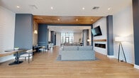Located at 3788 Old Port Royal Rd N in Spring Hill, TN, this thoughtfully designed single-floor space offers a generous layout encompassing a living room, dining room, and a dedicated office. The interior is characterized by light hardwood flooring throughout the main areas, soaring ceilings, and a neutral color palette of crisp whites and soft grays that creates a bright, airy atmosphere.

The living room is a welcoming area filled with natural light from large windows fitted with white blinds, providing views of the surrounding neighborhood. The space accommodates comfortable seating arrangements and is accented with contemporary artwork and warm accent tones.

The private office is defined by striking floor-to-ceiling glass partition walls with frosted logo detailing, offering both visibility and privacy. The office features a sleek work desk and additional seating, making it well-suited for professional use.

The dining room is carpeted and features a round glass-top table with mid-century modern wood chairs, a wall-mounted television, and a window that allows natural light to filter in. Colorful abstract artwork adds a creative touch to the space.

A larger open area connects the dining and living zones, highlighted by bold navy ceiling beams, recessed lighting, a fireplace, and expansive floor-to-ceiling windows that draw in abundant light and outdoor views, completing this polished and functional space.